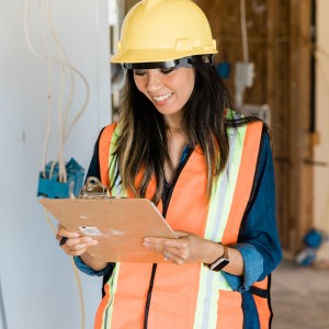 Image of a construction worker with a yellow hardhat, a blue, long-sleeved shirt, and a neon orange and yellow vest. They have long dark hair and tanned skin. They are standing in a room that is mid-renovation.